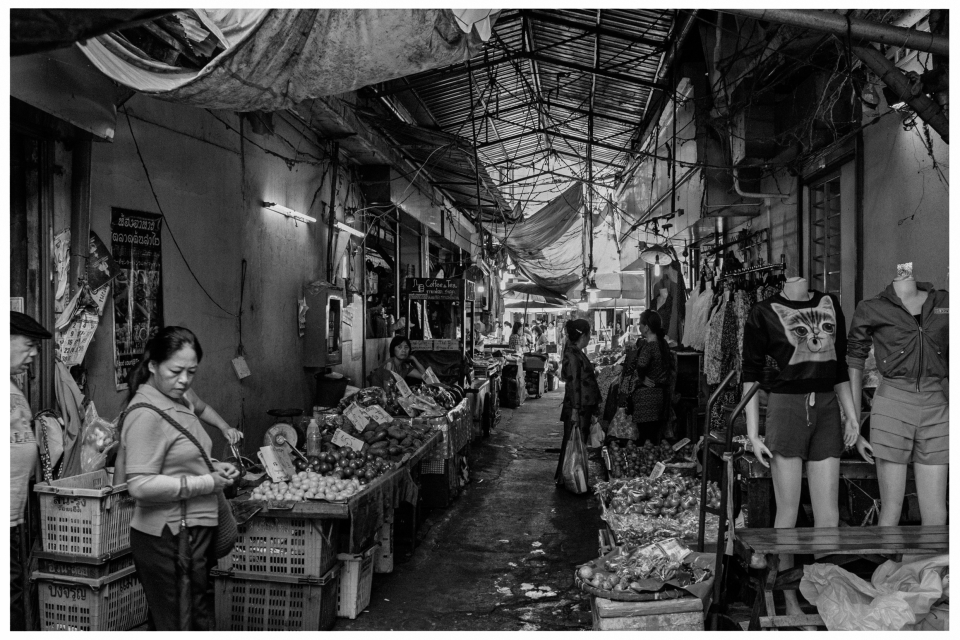 Überdachter Marktgang mit Obst, Gemüse und Kleidung, Menschen bewegen sich durch den Bereich.