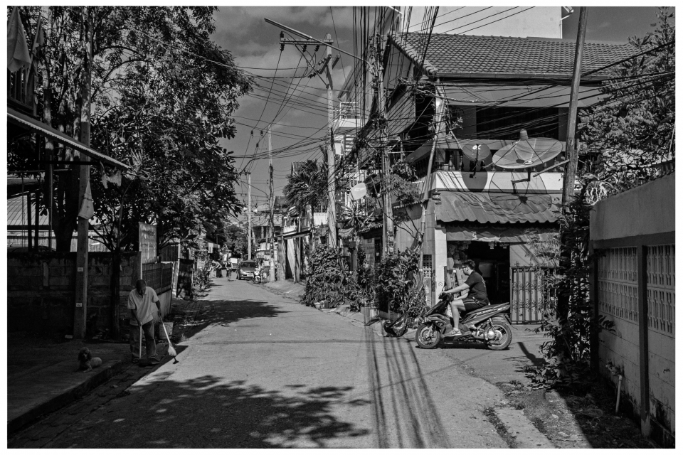 Städtische Wohnstraße mit Gebäuden, Satellitenschüsseln, Fußgängern und einem Motorroller.