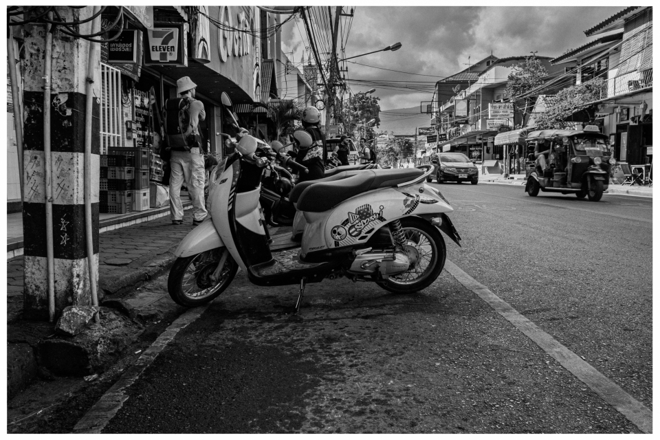 Straßenszene mit Motorrad, Personen vor Geschäft, Autos und Tuk-Tuk auf urbaner Straße.