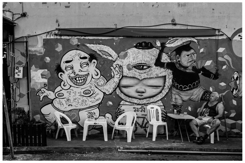 Straßenszene mit Graffiti von drei Figuren, Plastikstühlen und sitzender Person am Tisch.