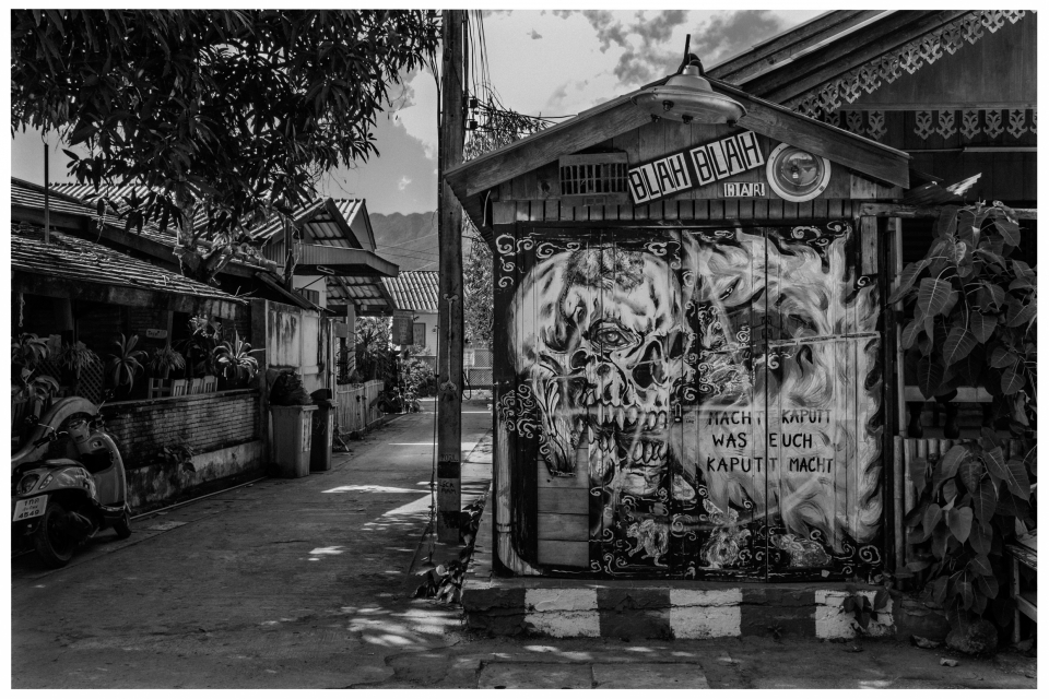 Schwarz-weiße Gasse mit bemalter Hauswand, Skelettmotiv und Schriftzug, angrenzender Motorroller.