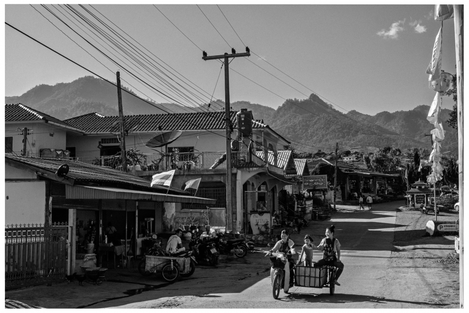 Dorfstraße mit Wohnhäusern, Geschäften, Familie auf Motorrad und Berge im Hintergrund.