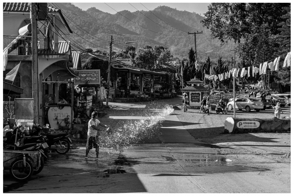 Eine Person spritzt Wasser auf eine Straße, umgeben von Gebäuden, Fahrzeugen und bewaldeten Hügeln.