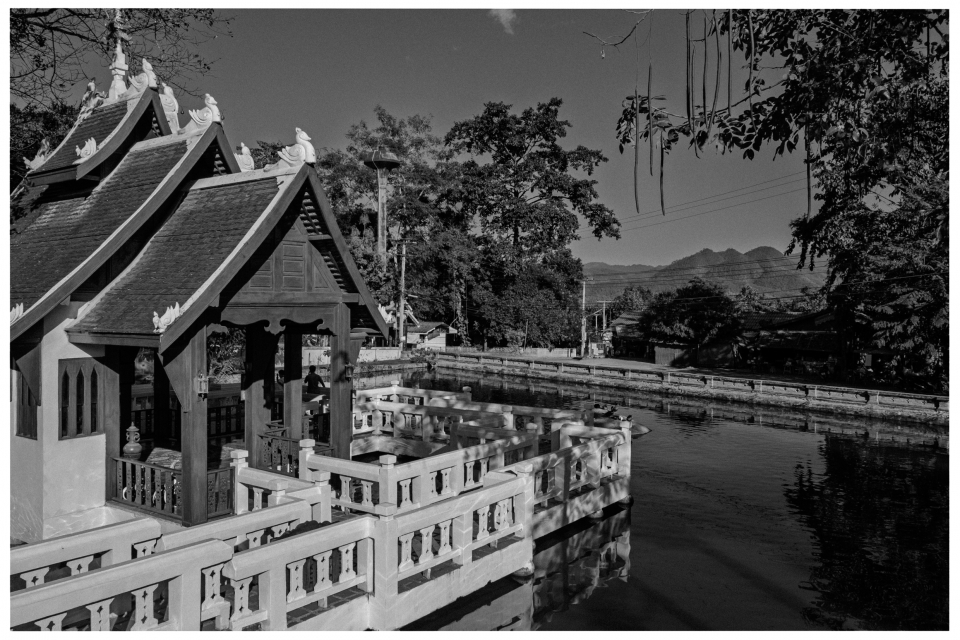 Traditionelles Gebäude am Flussufer, umgeben von Bäumen, Bergen im Hintergrund.