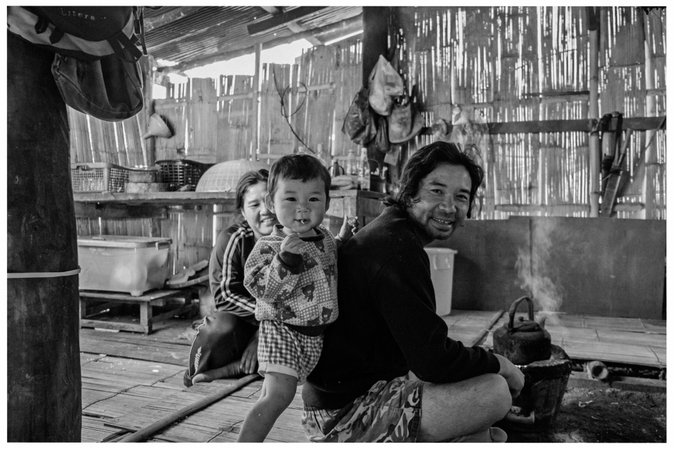 Gruppe in Bambusgebäude, Mann sitzt vor rauchendem Topf, Kind schaut zur Kamera.