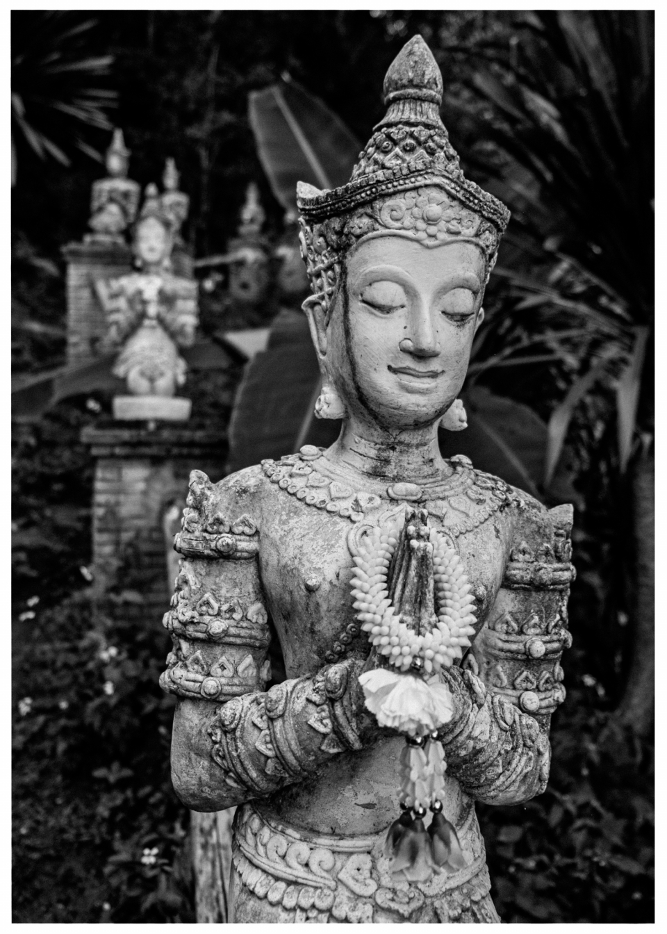 Steinerne Statue in Gebetspose mit weiteren ähnlichen Skulpturen im grünen Hintergrund.