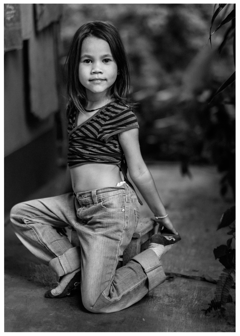 Mädchen in gestreiftem Oberteil und Jeans kniet in unscharfem Innenraum, Schwarzweißfoto.