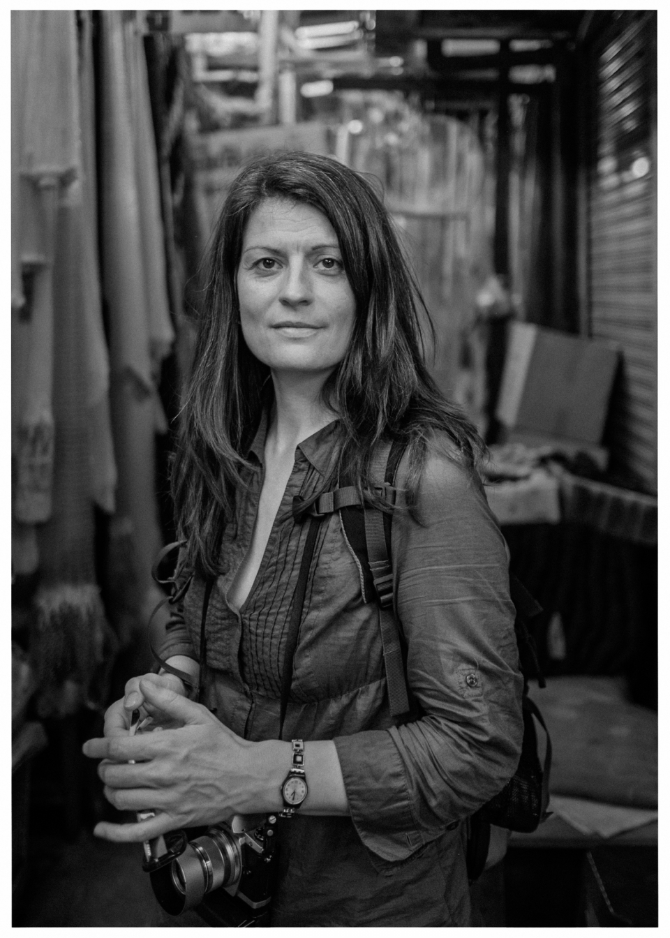 Frau mit Kamera und Jacke in Innenraum, umgeben von Textilien und Gegenständen.