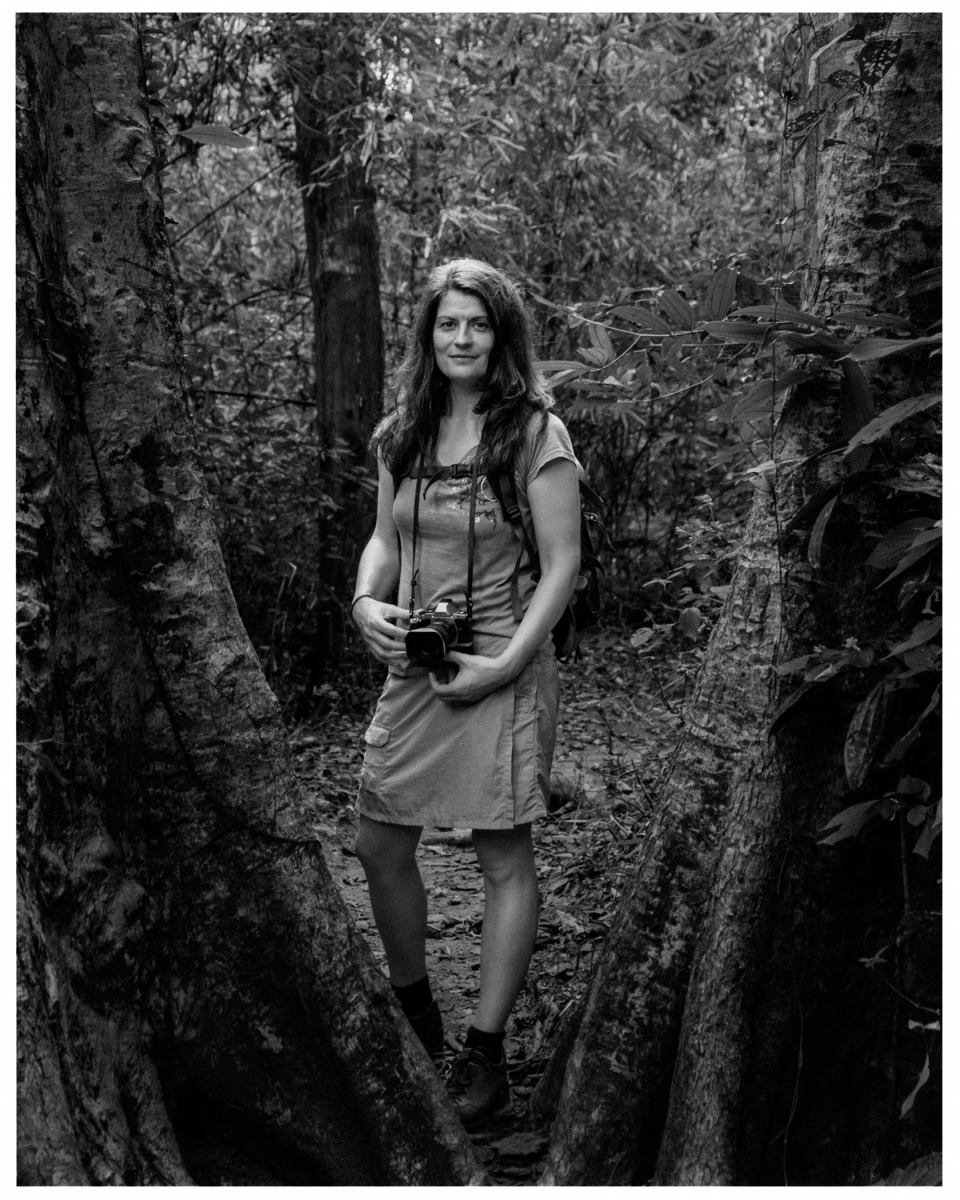 Frau im Wald, umgeben von Bäumen, hält Kamera in den Händen.